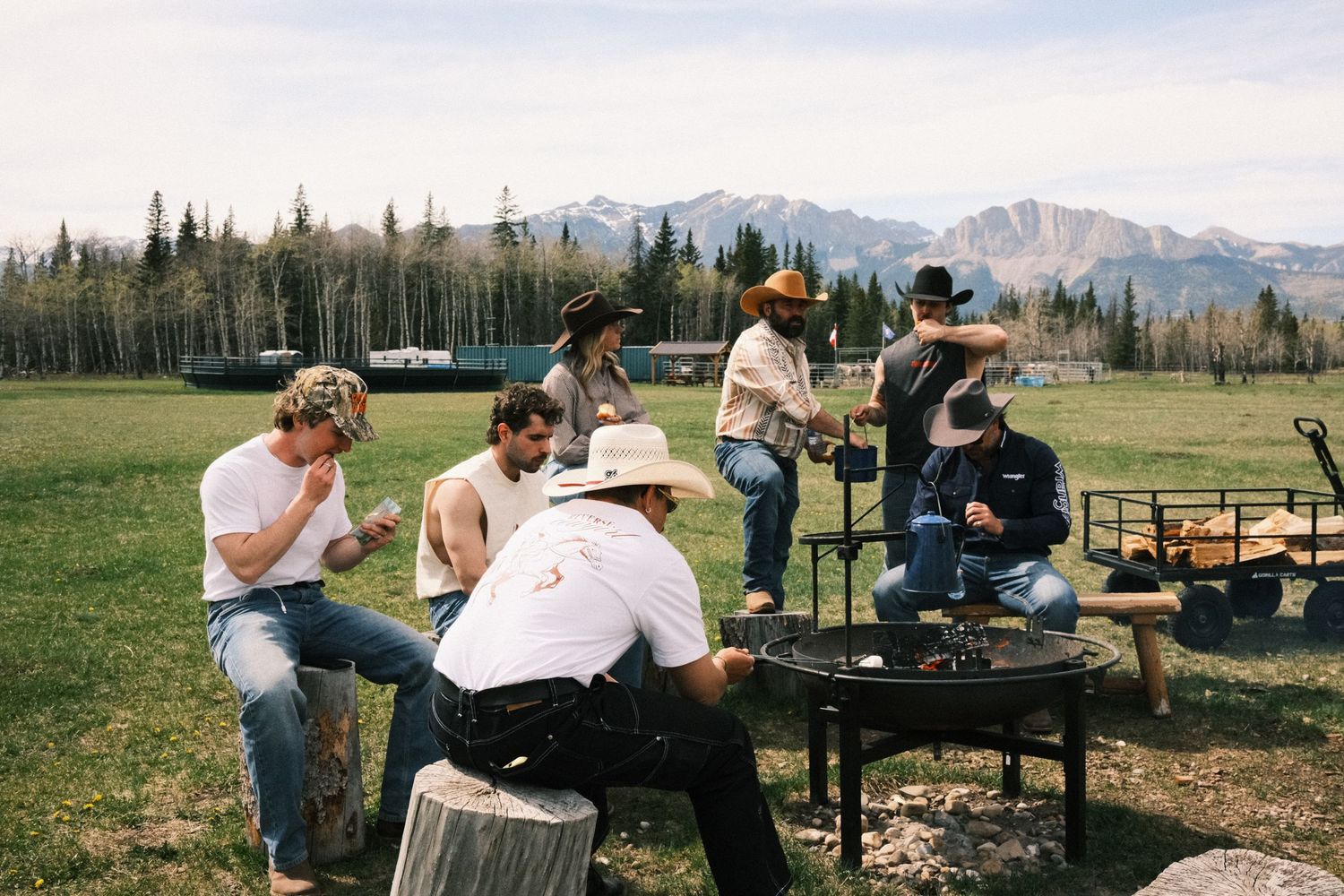Group of friends gathered around a fire pit on a ranch with mountain views and green meadows.
