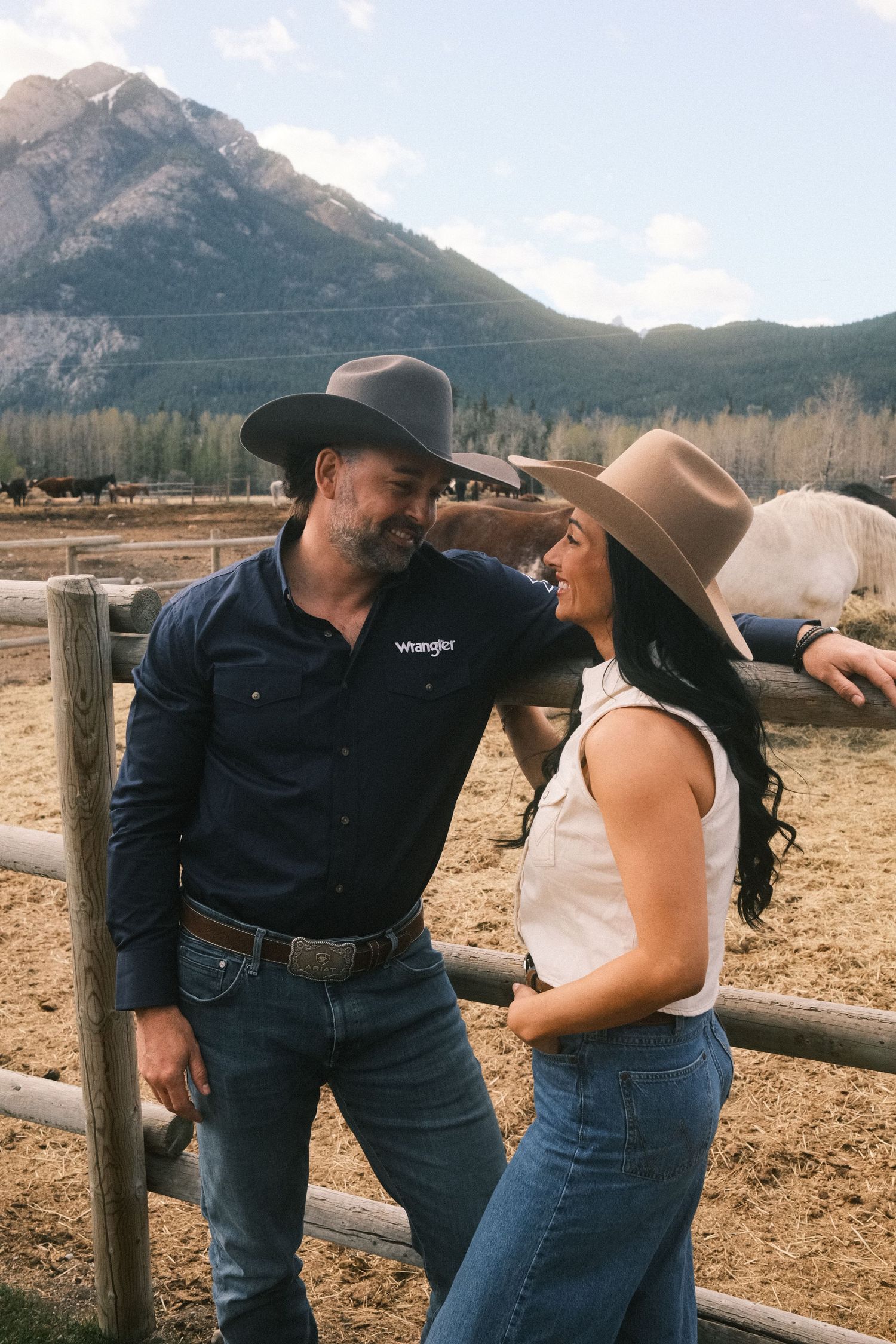 A couple in cowboy hats and Wrangler shirts smile at each other by a ranch fence with mountains in the background.