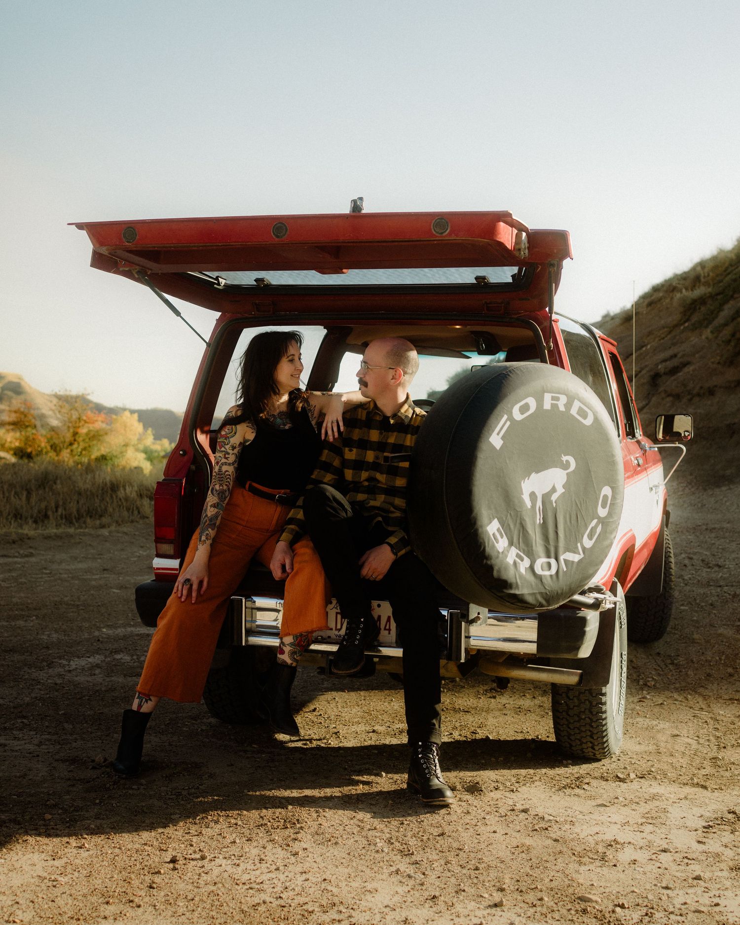 Couple posing playfully around a vintage red truck in a desert setting.