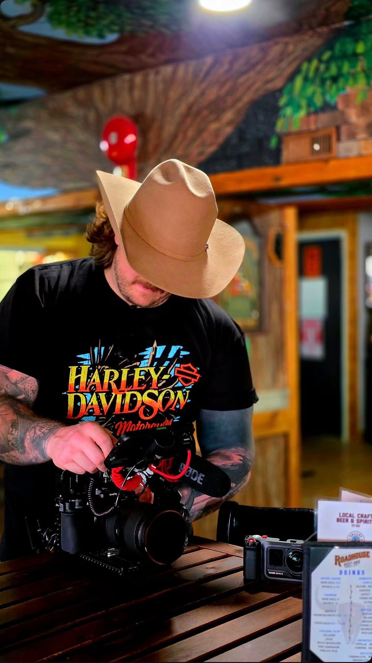 Tattooed man in cowboy hat and Harley-Davidson shirt operating a professional camera in a rustic bar setting.