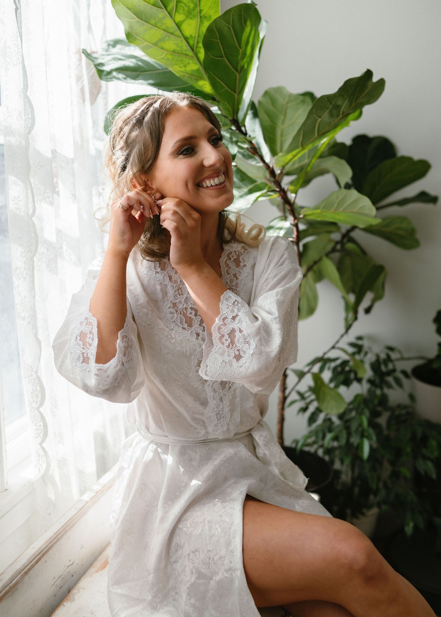 Woman in white lace robe posing by window with fiddle leaf fig plant.