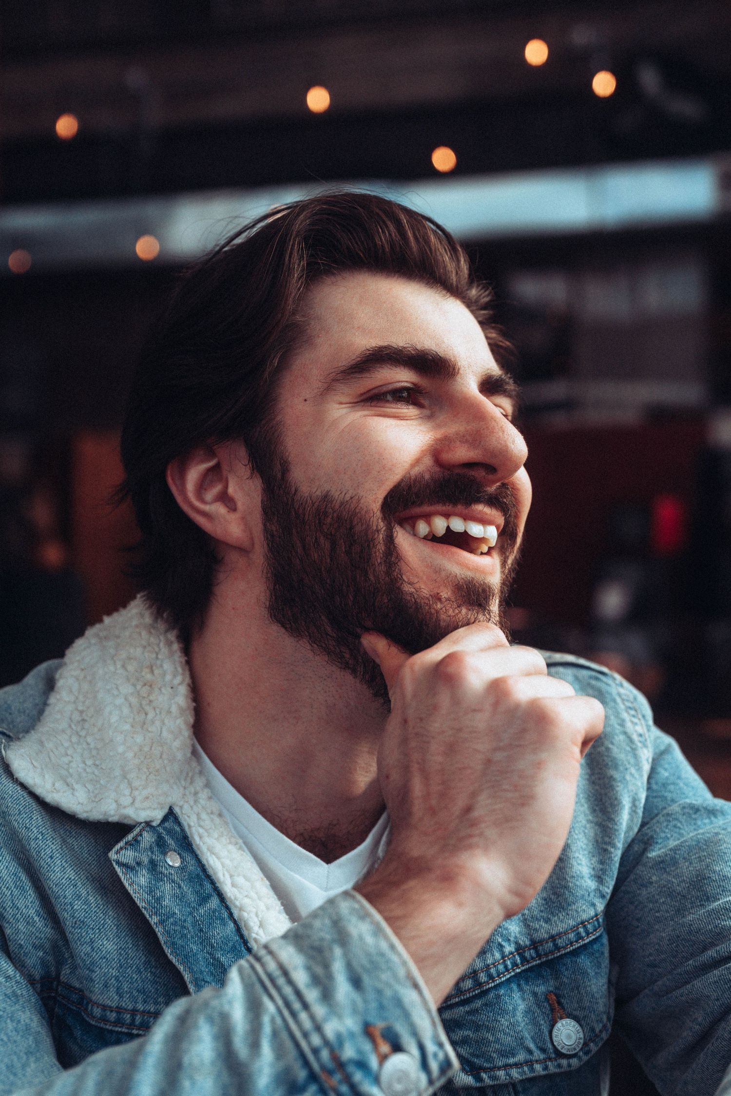 Person in denim jacket smiling warmly in atmospheric bar lighting.