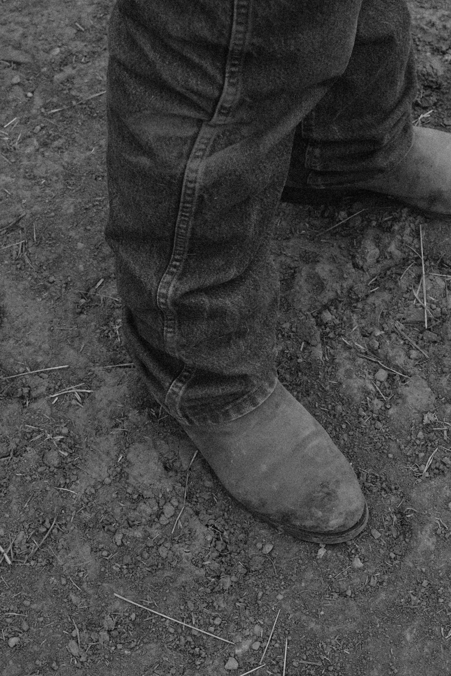 Black and white photo of worn cowboy boots and jeans standing on muddy dirt ground.