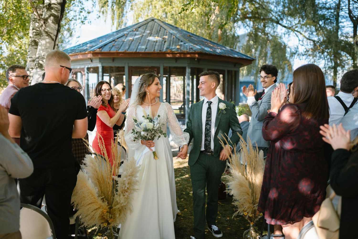 Outdoor wedding celebration with guests and natural decor under willow trees.