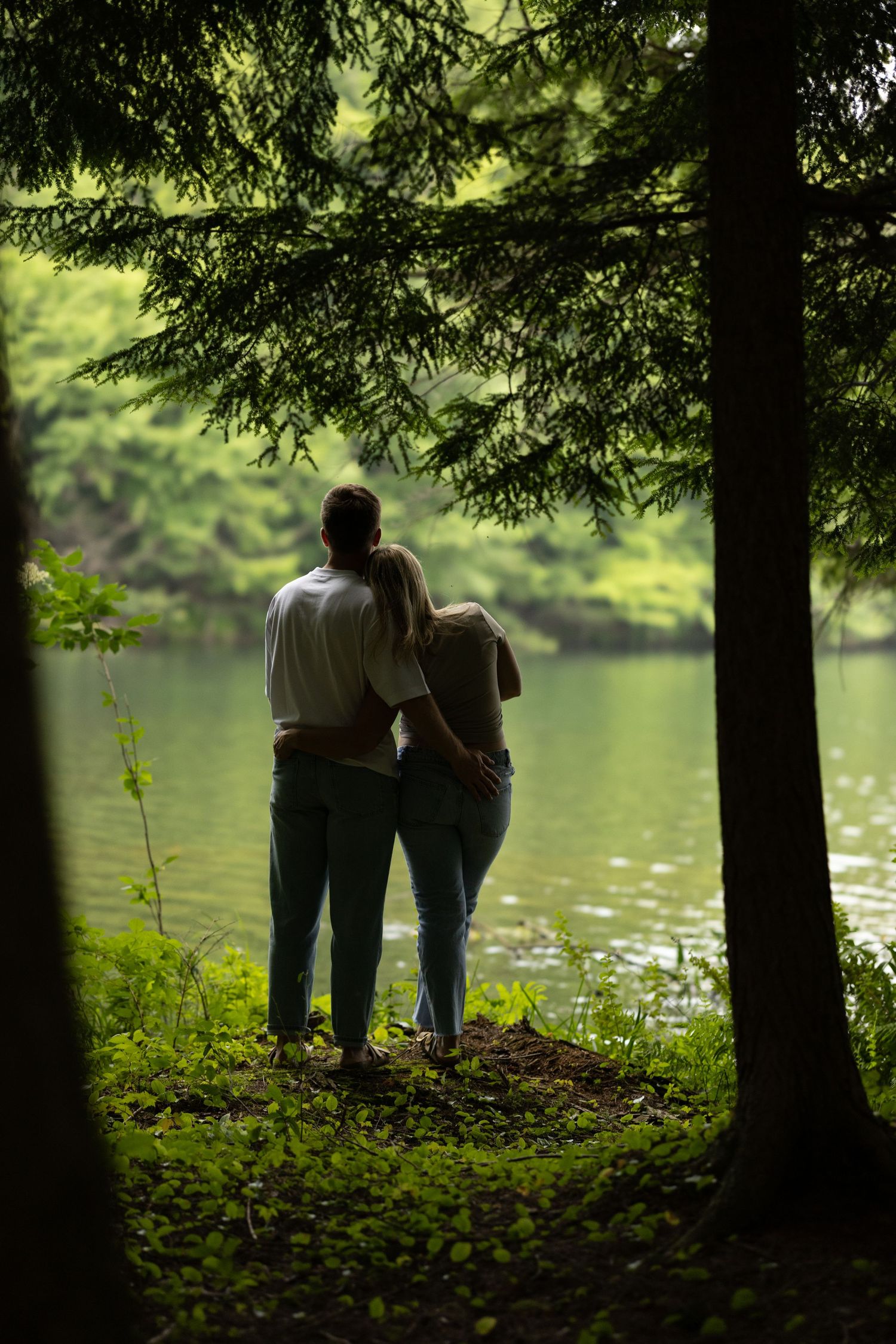 Silhouetted couple standing by tranquil lake under tree canopy.