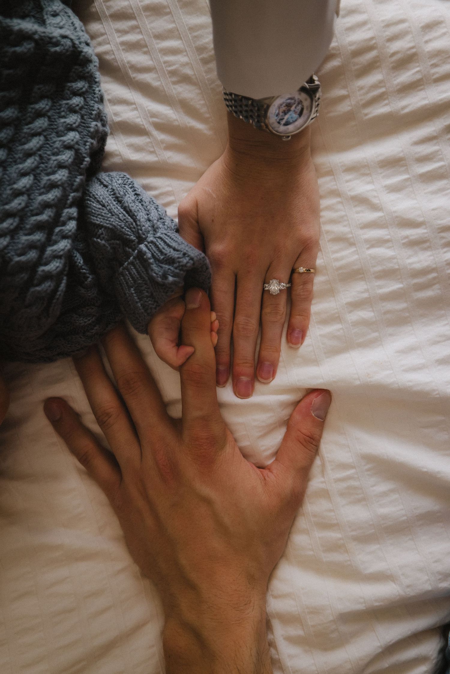 Hands with engagement ring rest on white bedding, showcasing intimate family moment.