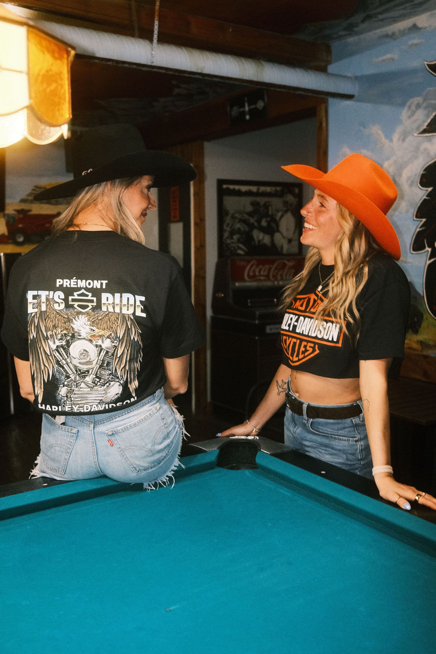Two women in Harley-Davidson tees and cowboy hats chat by a pool table in a rustic bar setting.