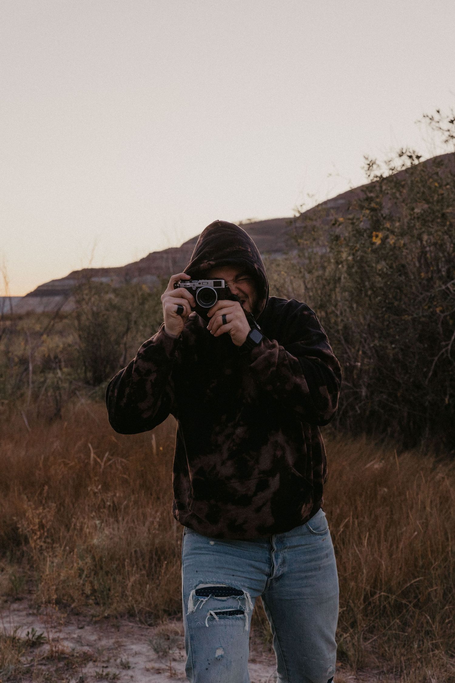 Person in dark hoodie and ripped jeans photographing with vintage camera outdoors at dusk near rocky hills.