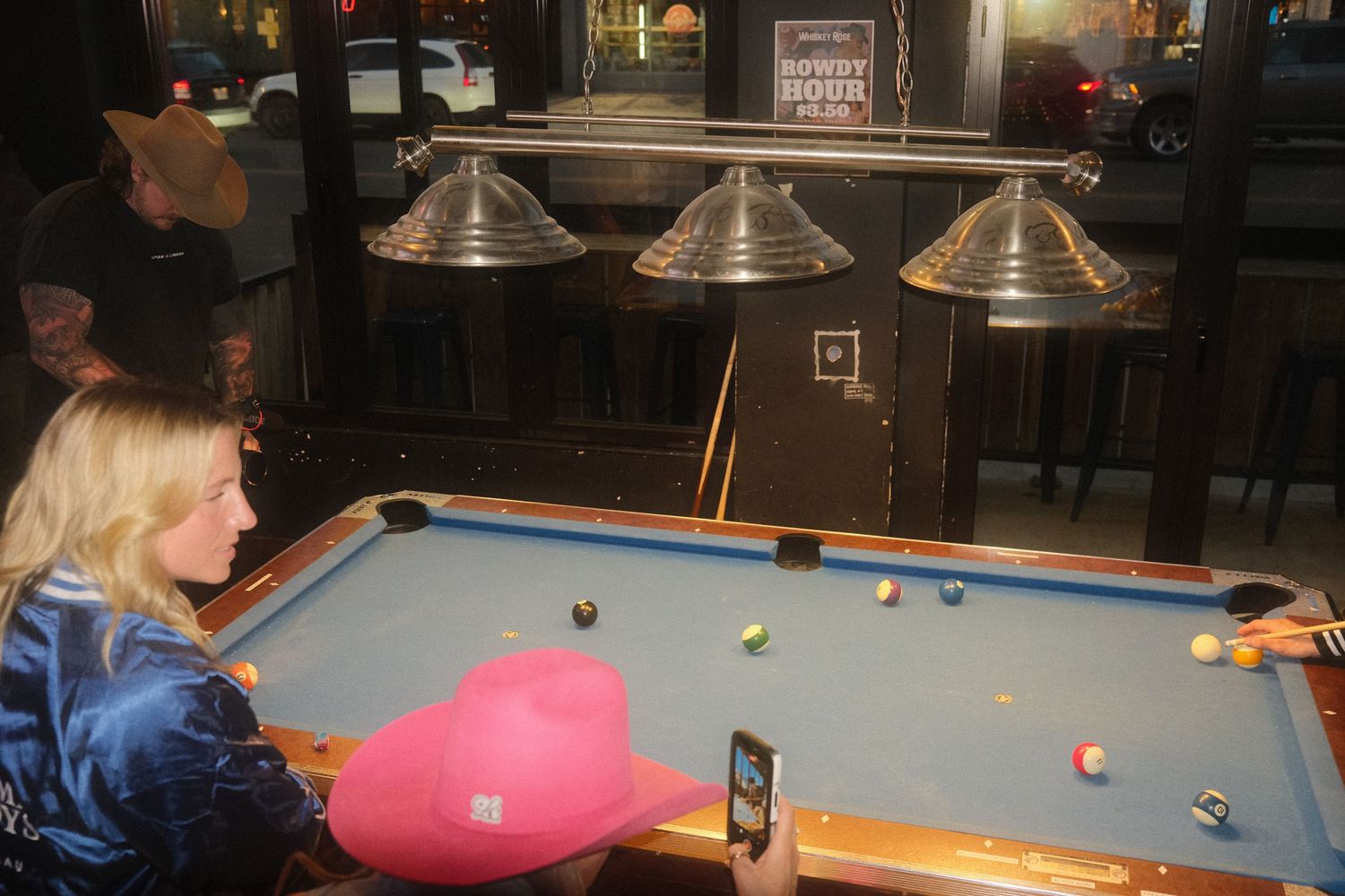 Woman in blue jacket playing pool at a bar with pink hat on table and billiard balls on blue felt table.