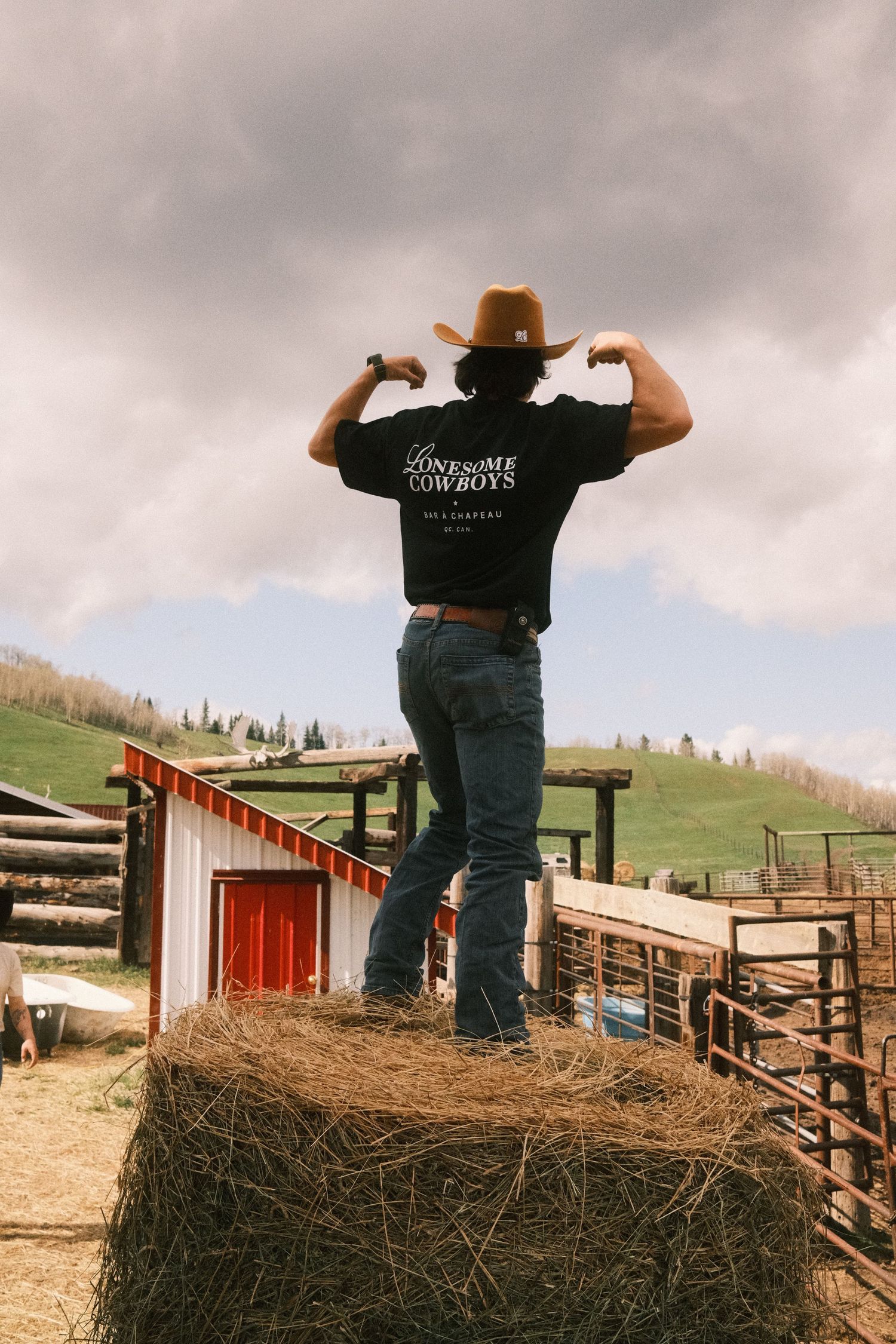 A cowboy flexing muscles atop a hay bale at a ranch, wearing a black Lonesome Cowboys tee and brown hat.