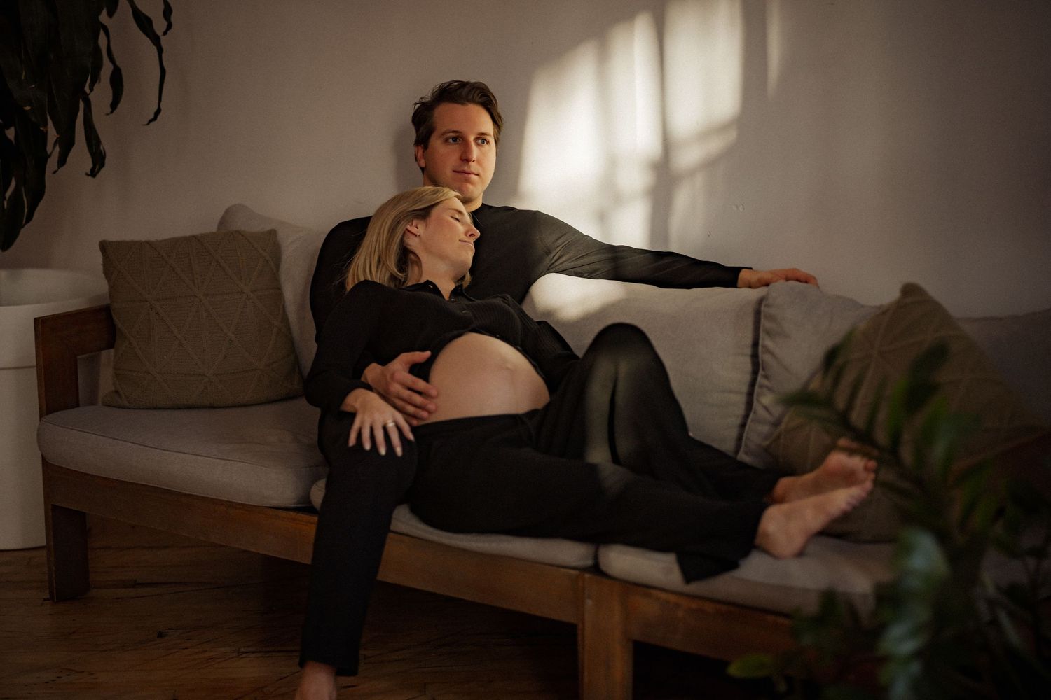 Couple relaxing together on a gray couch in a sunlit room with plants.