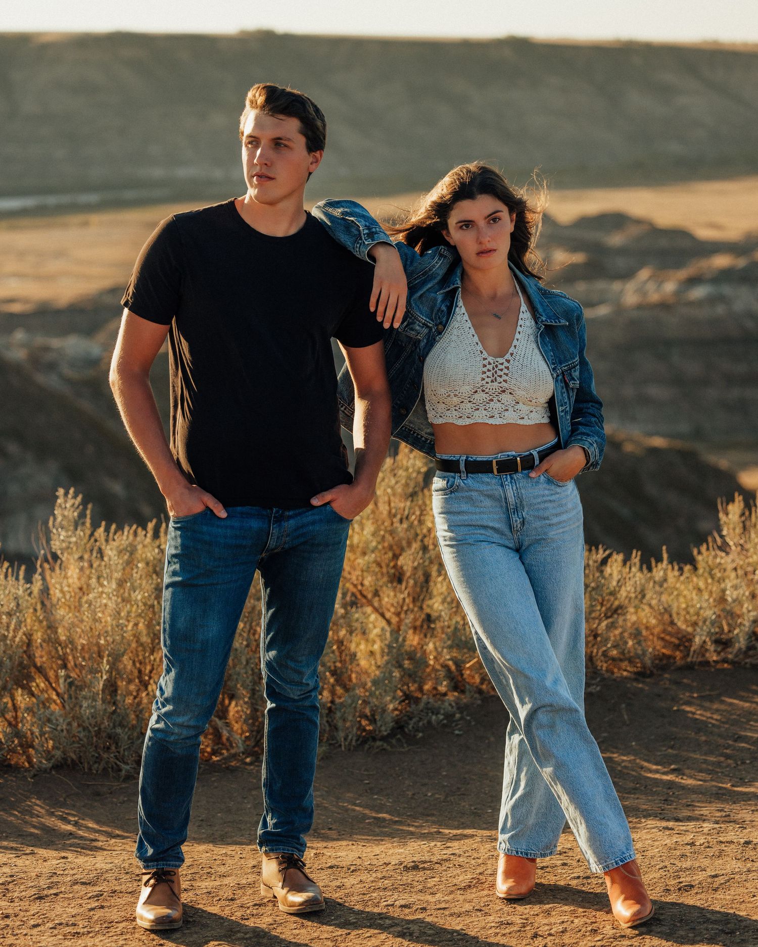 Stylish couple in denim and black posing against desert landscape at sunset.