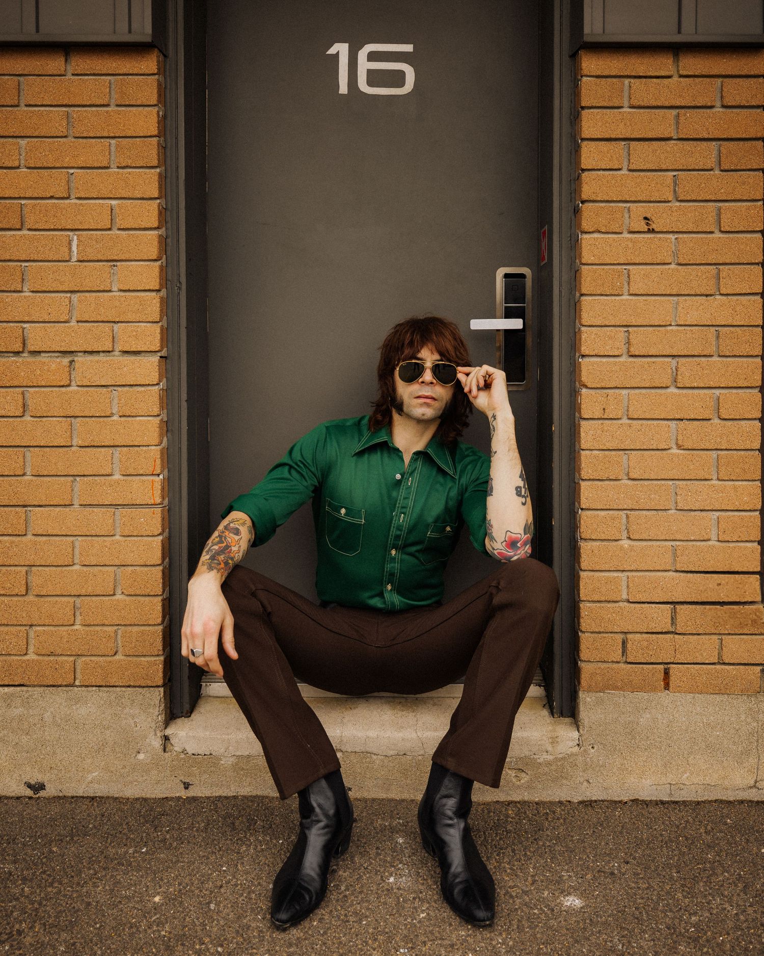 Person in green shirt and dark pants sitting against brick wall with sunglasses.
