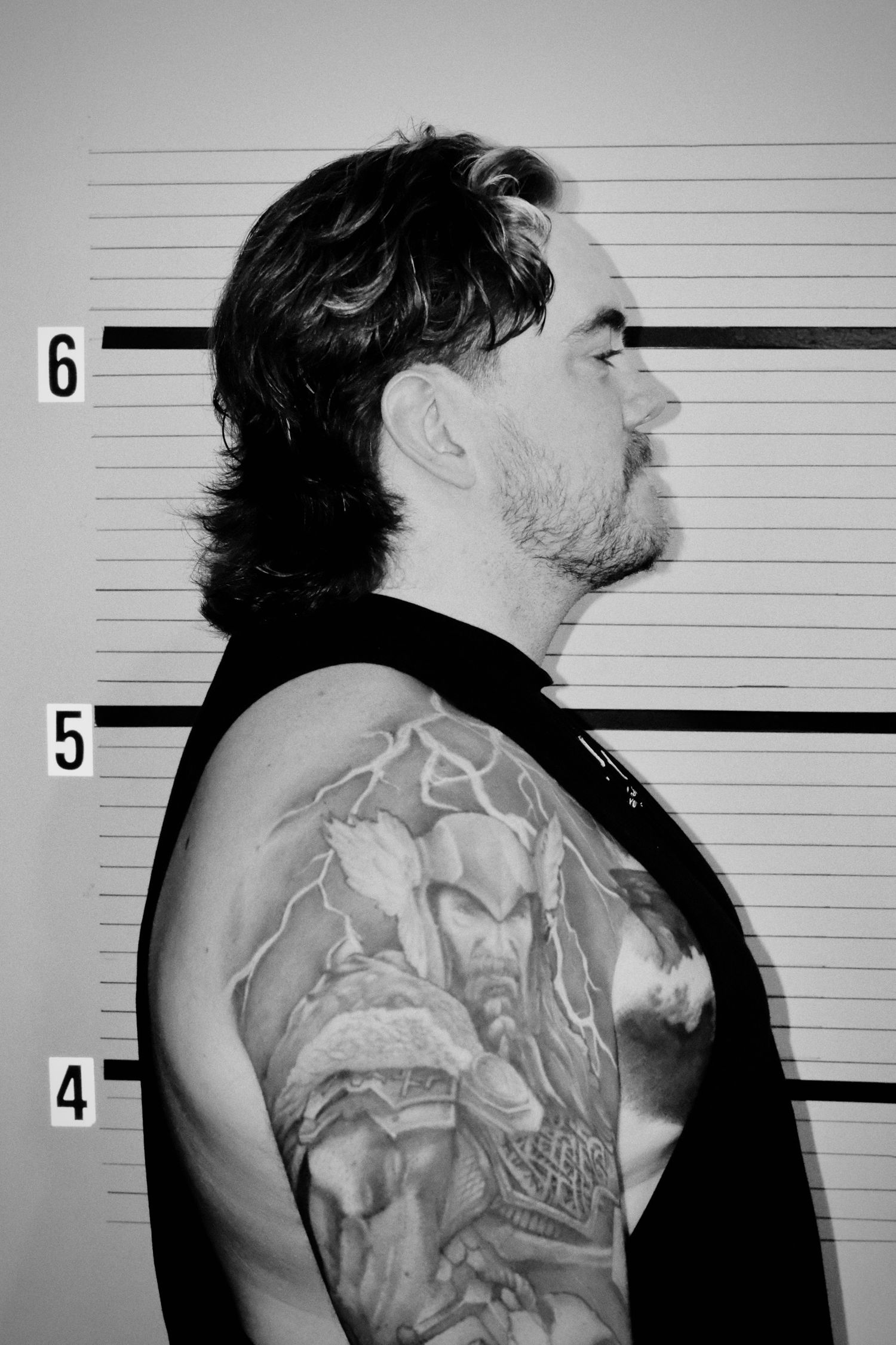 Black and white mugshot of a tattooed man with a mullet hairstyle standing against a height chart wall.