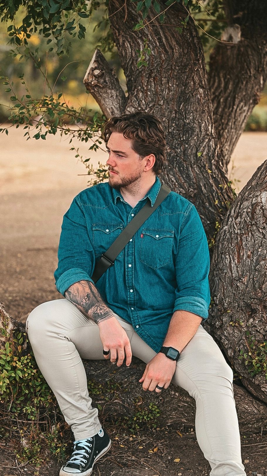 Man in teal Levi's shirt with tattoos sits against a tree outdoors, wearing khakis and Converse sneakers.