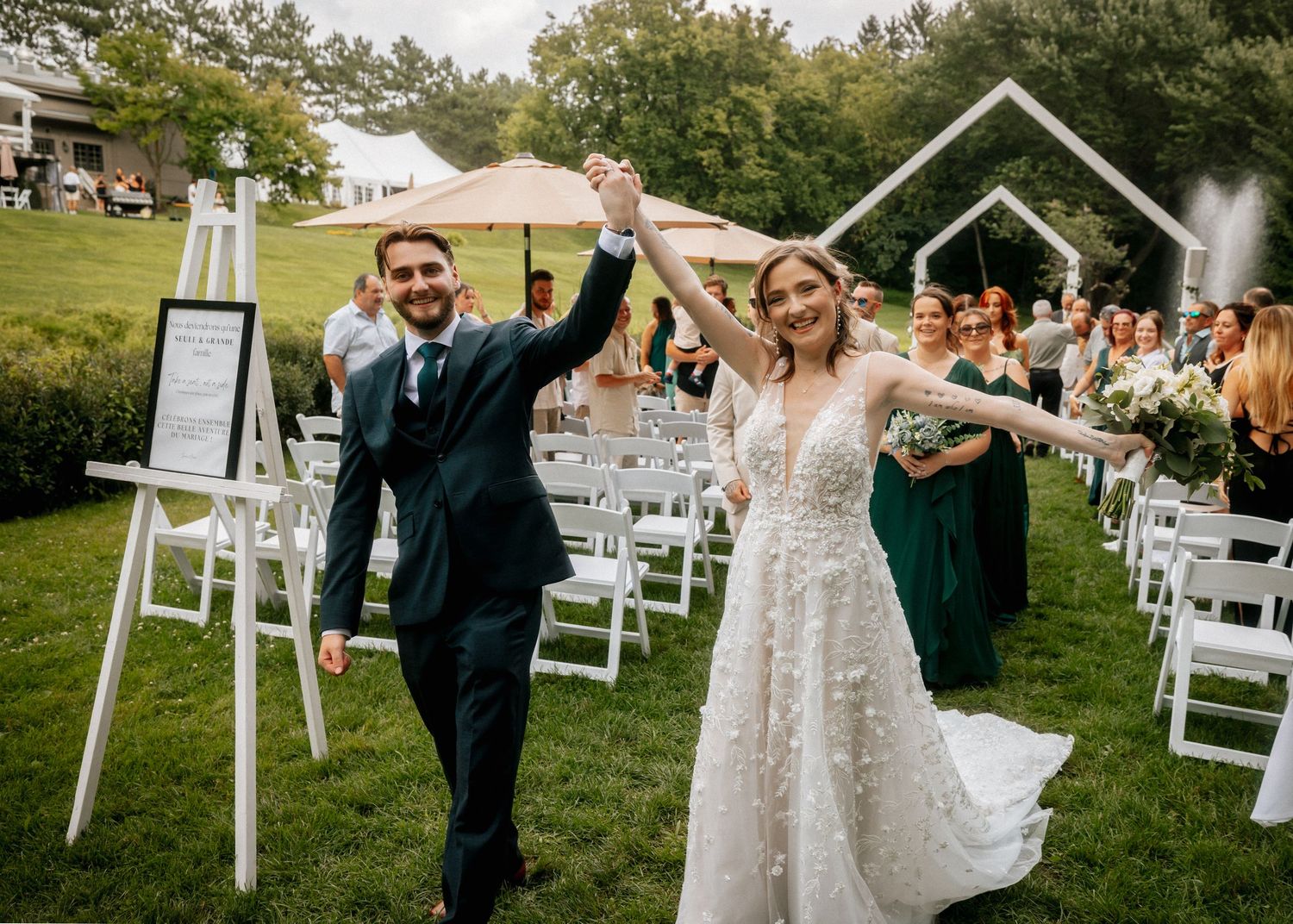 A wedding couple celebrates with guests during an outdoor ceremony with white chairs on a grassy lawn.