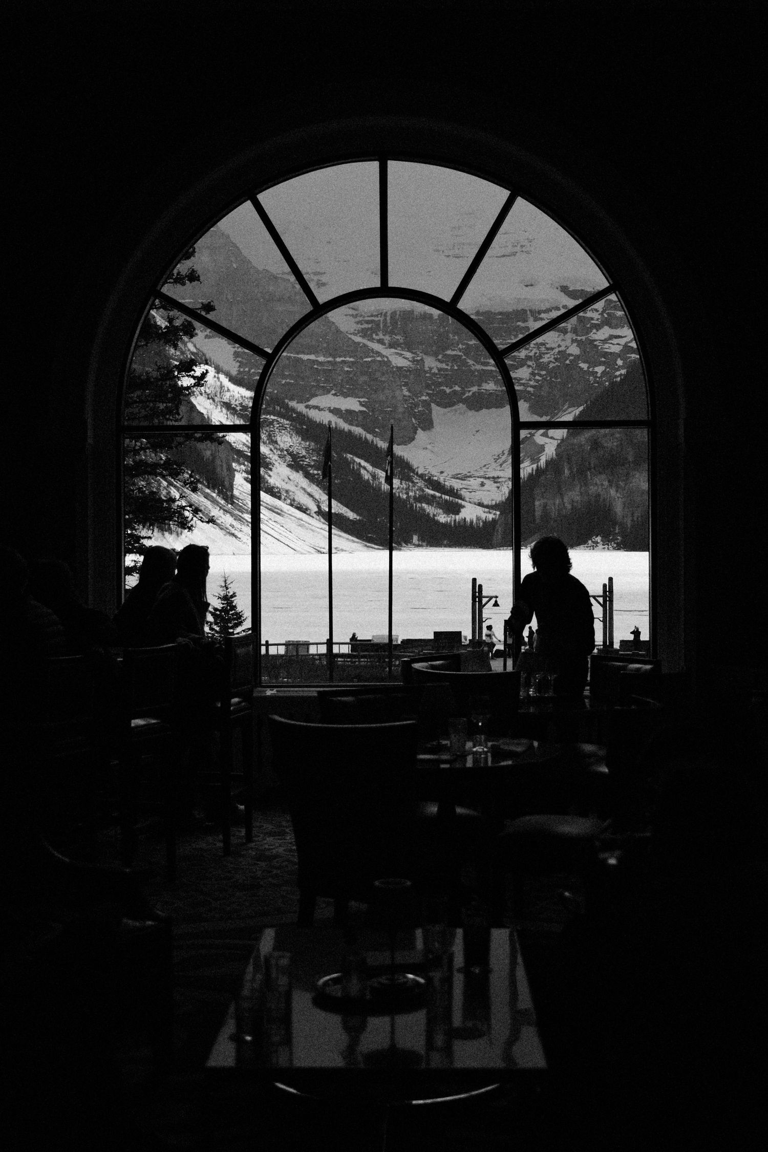 Silhouette of person standing before arched window overlooking snowy mountain lake landscape in black and white.