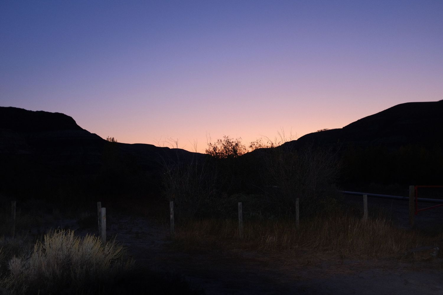 Twilight sunset silhouettes rugged desert mountains with a colorful pink and purple sky above a rural fenced road.