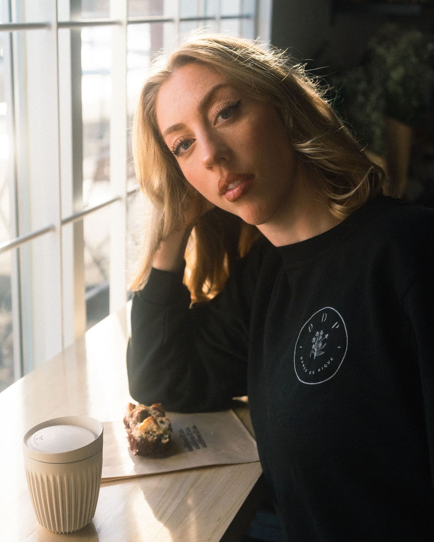 Figure in black sweater sits contemplatively by window with coffee cup.
