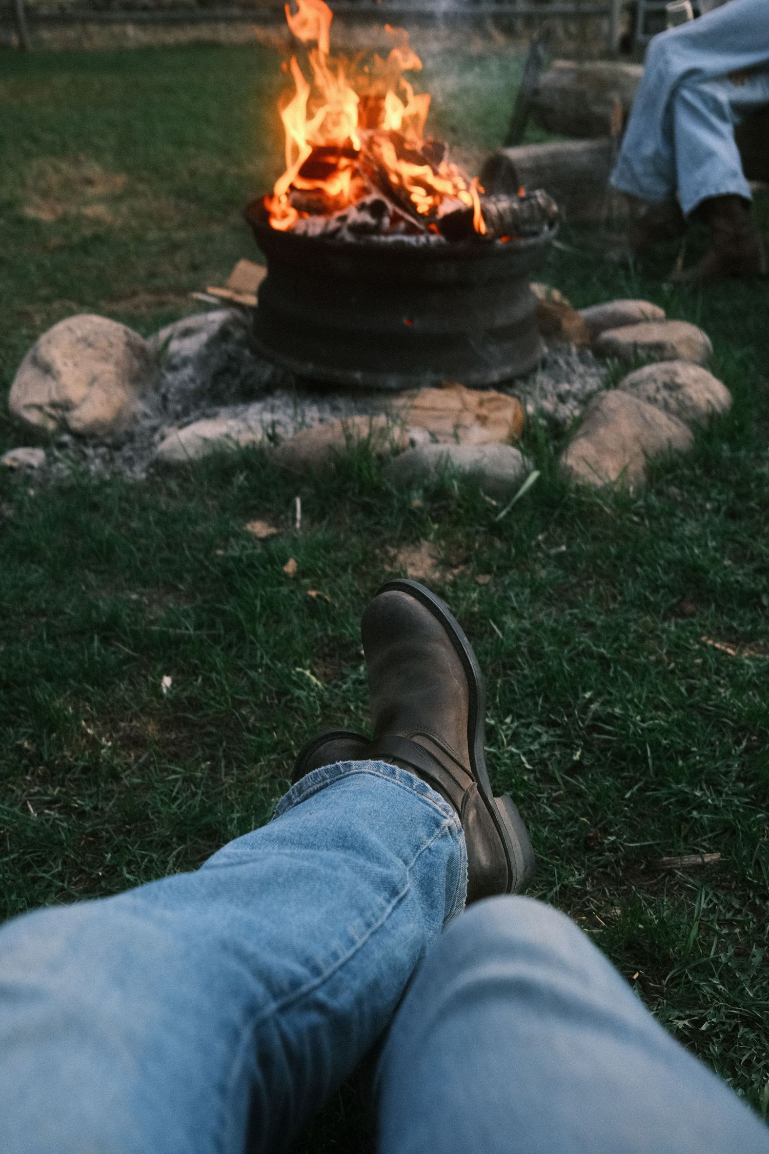Person in jeans and boots relaxing by a crackling campfire surrounded by rocks on green grass.