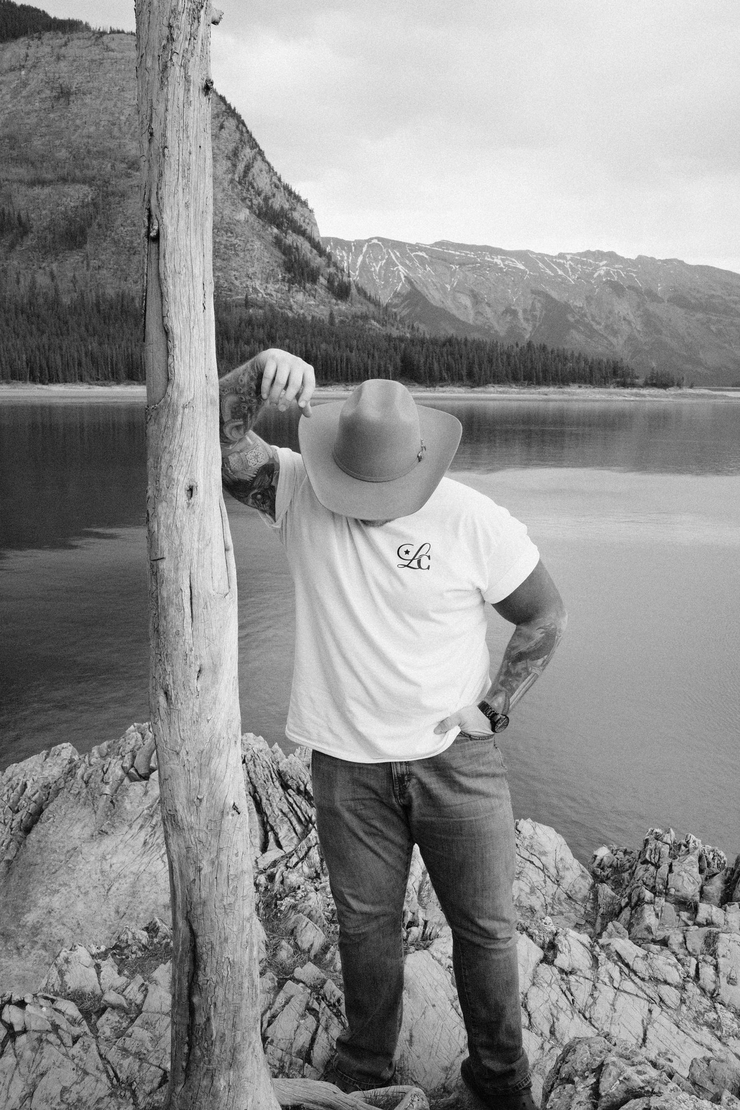 Man in cowboy hat and jeans leans on wooden post by mountain lake in black and white photo.