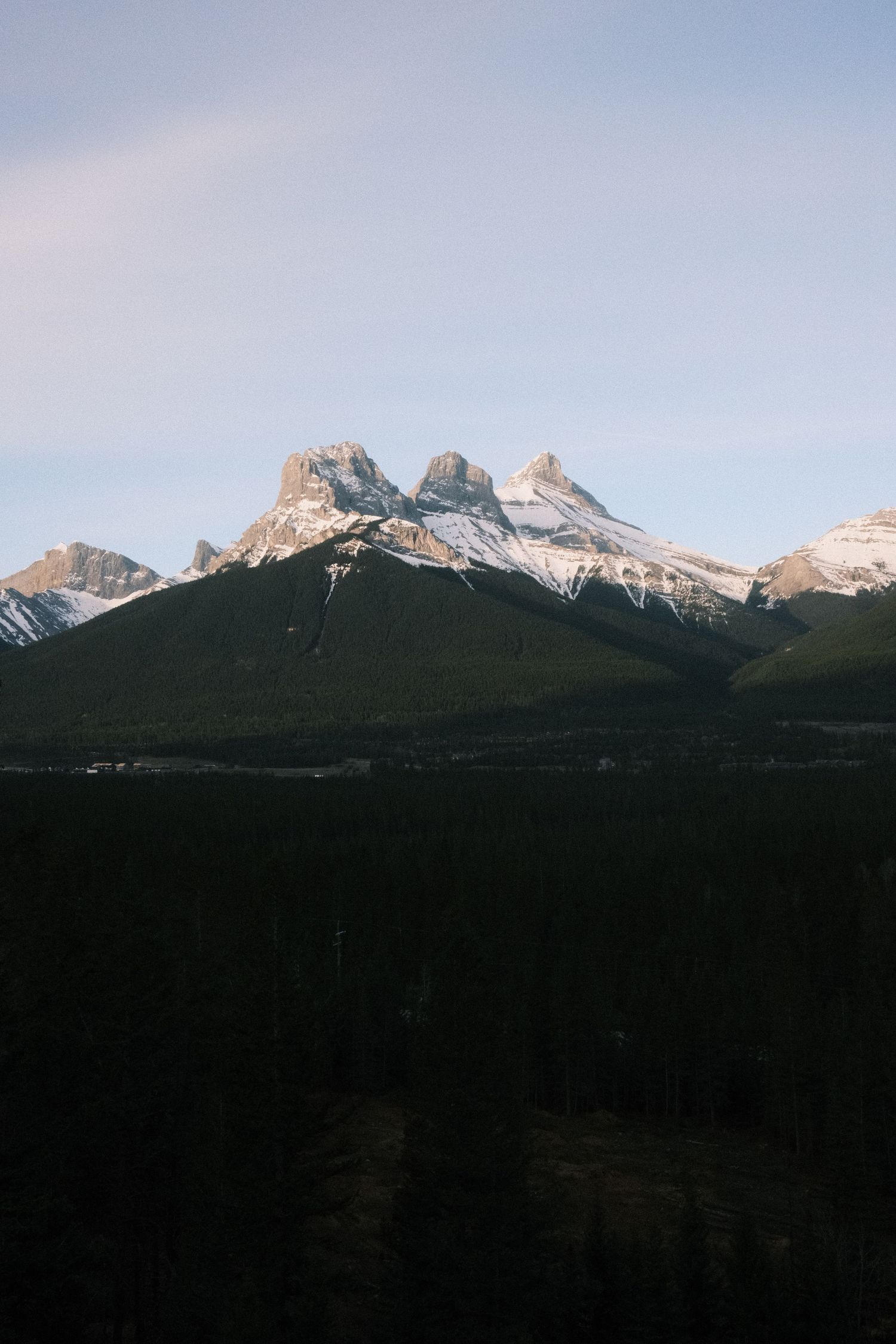 Snow-capped Three Sisters mountain peaks rise above dense evergreen forest in Canmore, Alberta, Canada.