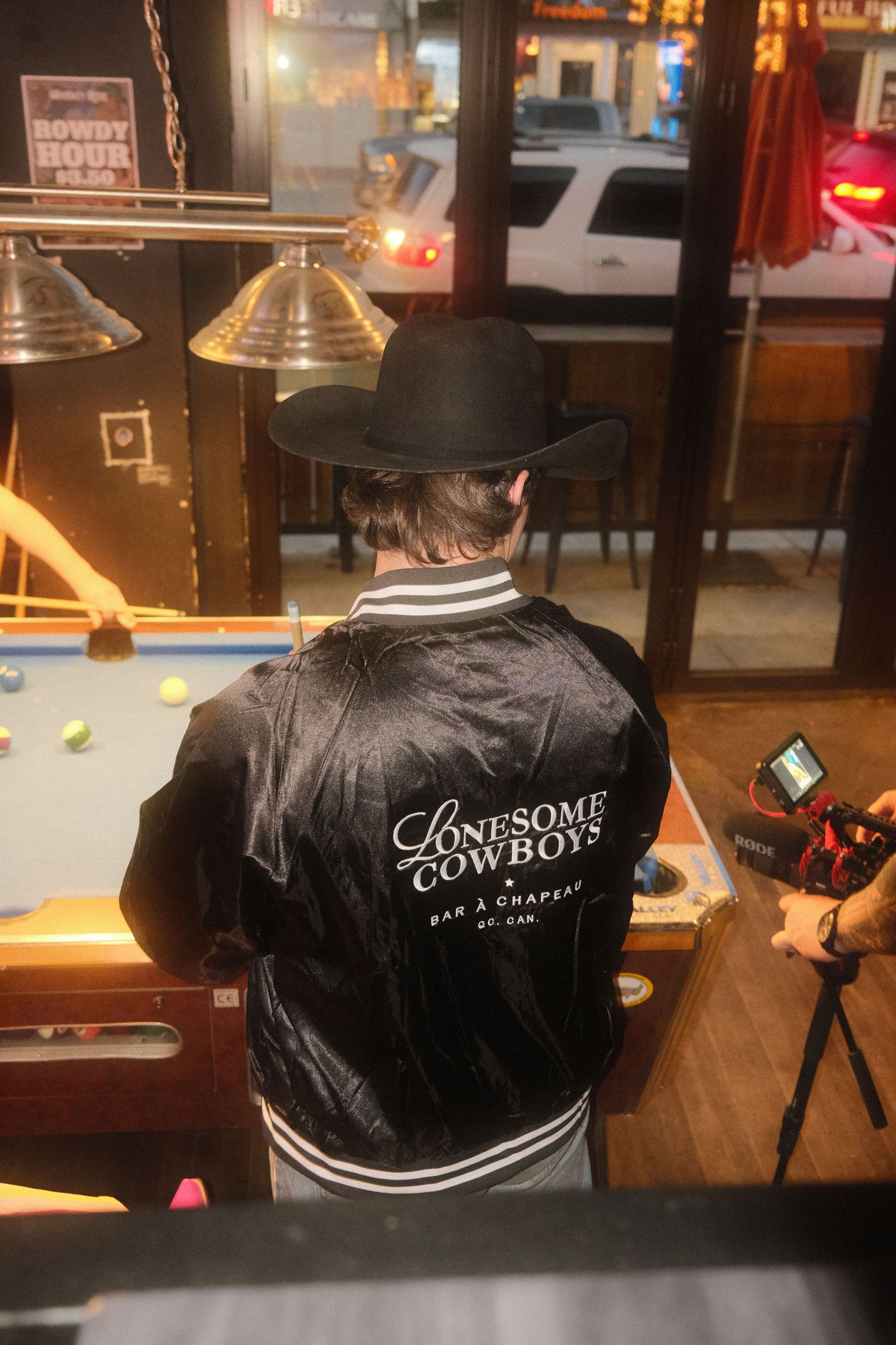 Man wearing black cowboy hat and Lonesome Cowboys bar jacket standing near pool table in western bar.