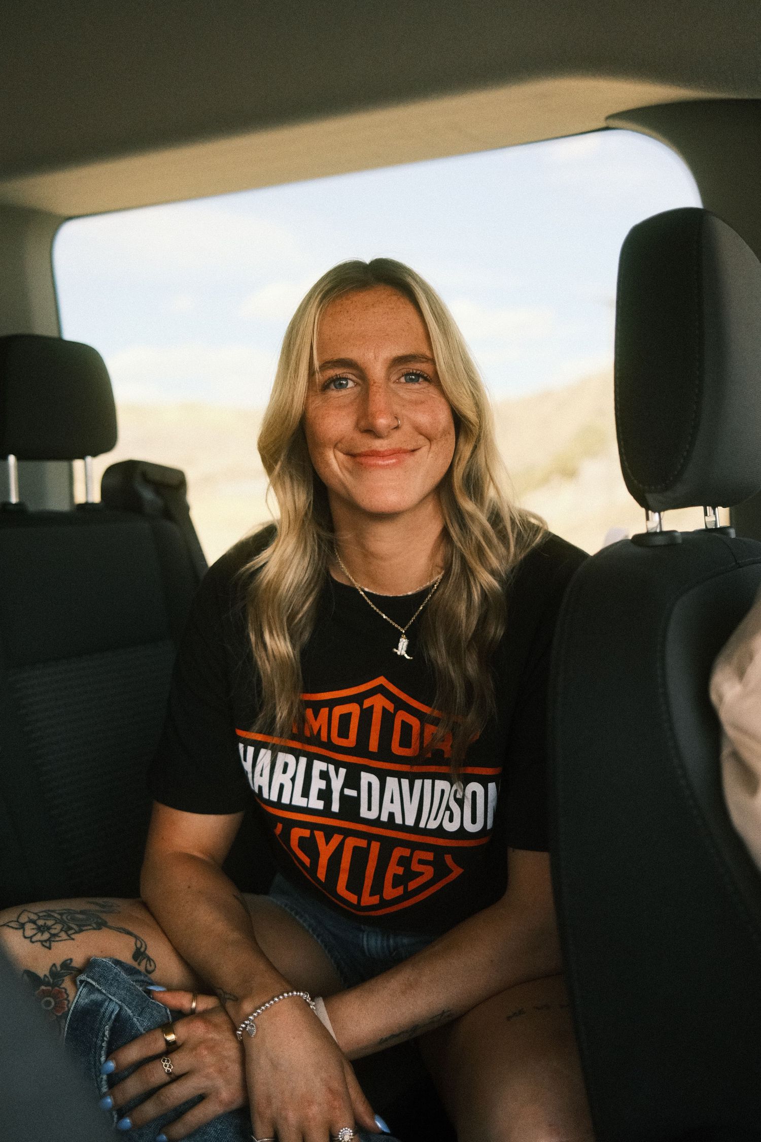 Smiling blonde woman in Harley-Davidson t-shirt sitting in back seat of vehicle with desert landscape behind her.