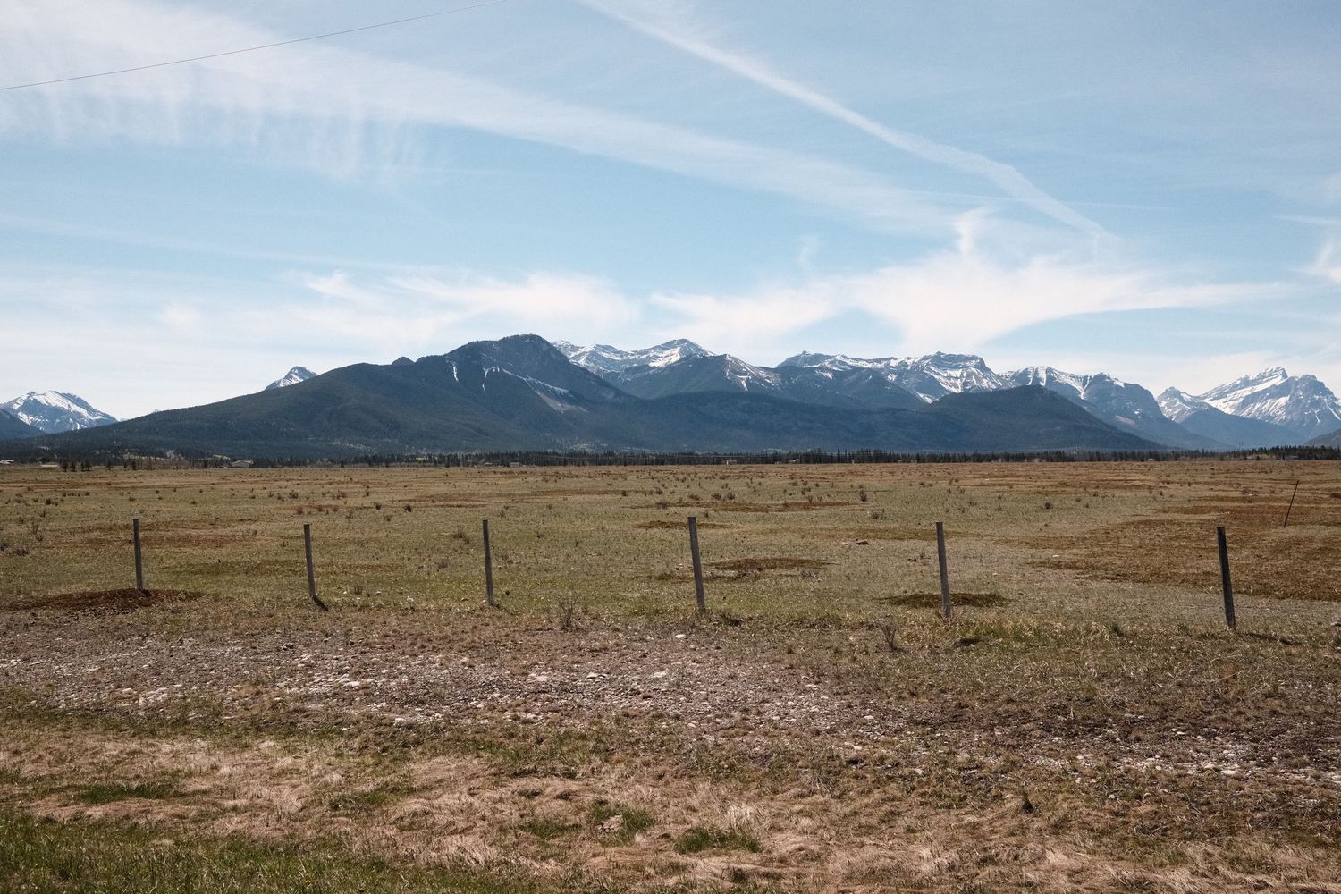 Snow-capped Rocky Mountains rise behind a vast open ranch field with rustic fence posts under a blue sky.