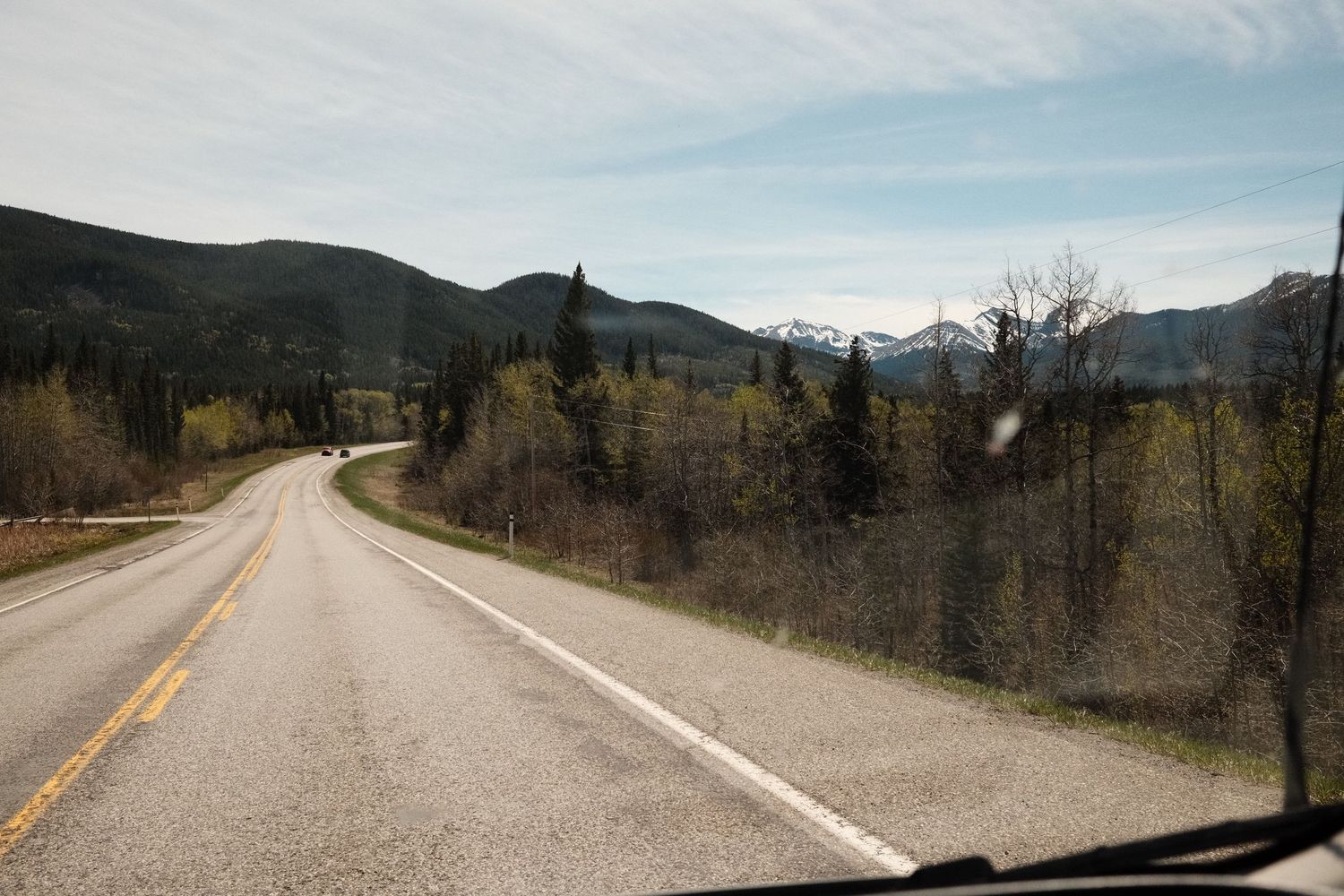 A scenic two-lane highway winds through lush green forests toward snow-capped mountains under a partly cloudy sky.