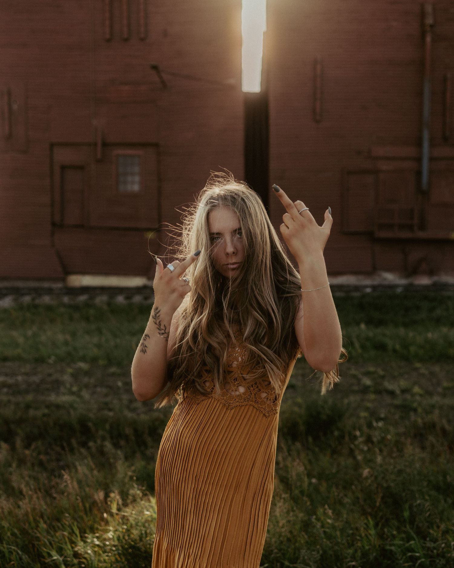 Woman in golden dress with tattoos poses outdoors near brick buildings with dramatic backlight sunset.