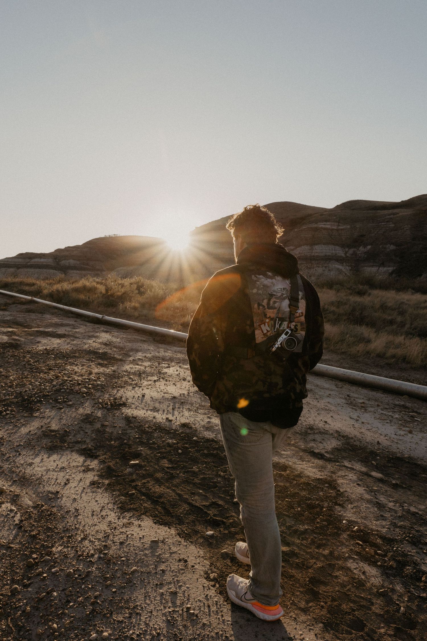 Person standing on a desert road at golden hour with sun rays shining behind rocky hills in the background.