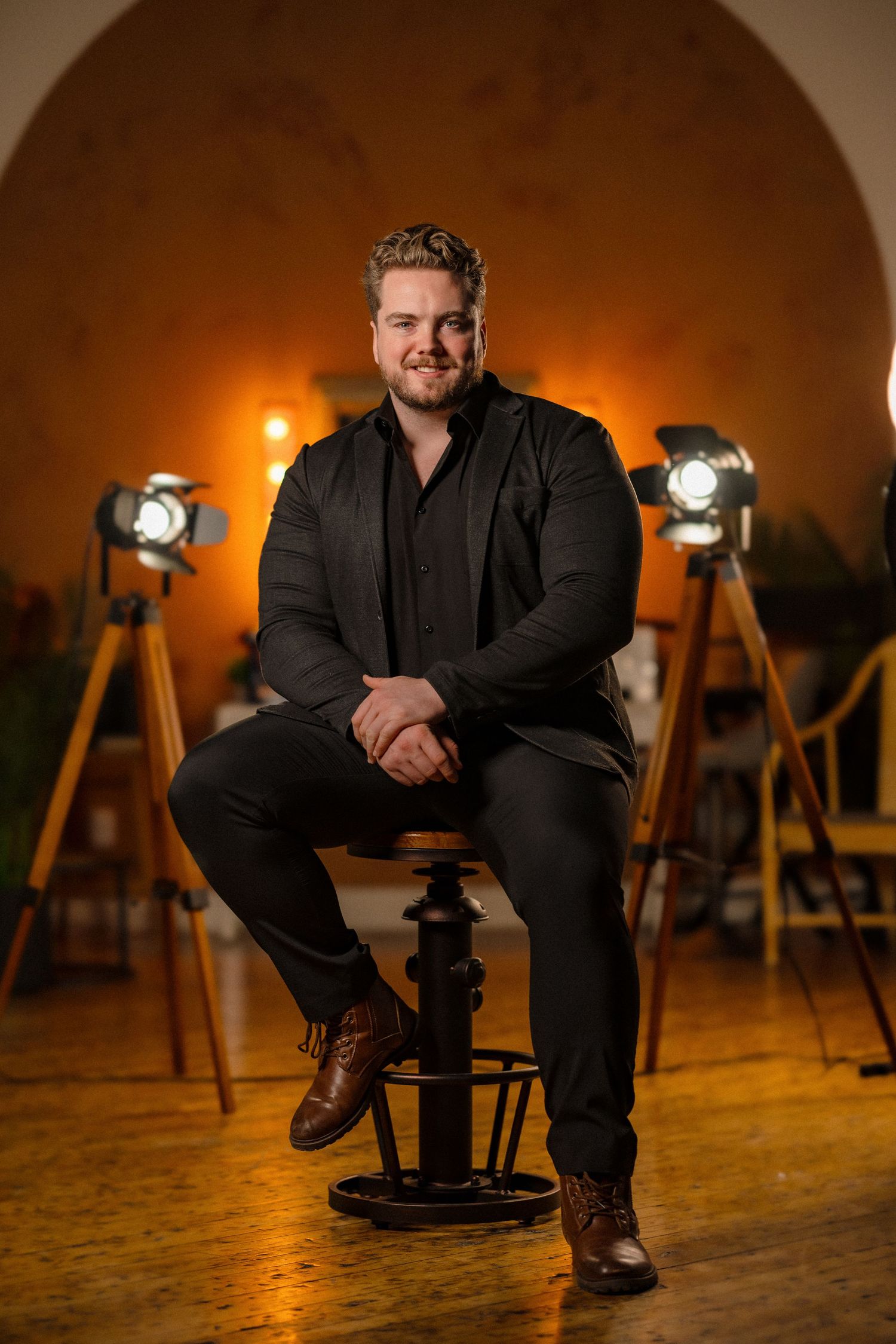 Person in all black outfit poses on stool with vintage spotlights in background.