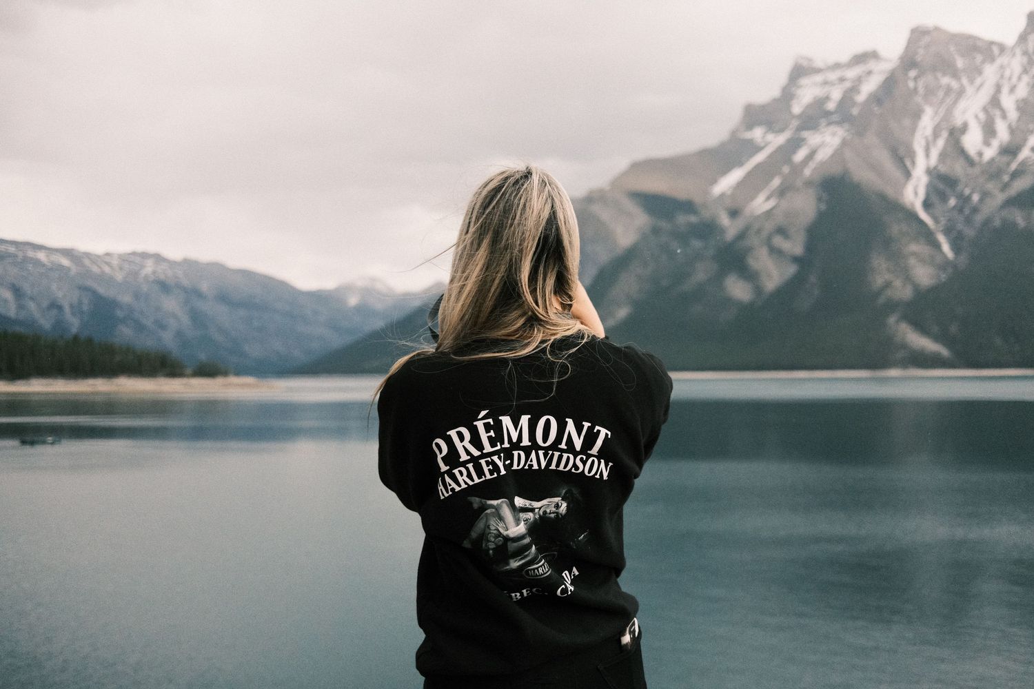 Woman in Prémont Harley-Davidson black sweatshirt overlooking a serene mountain lake with snow-capped peaks.