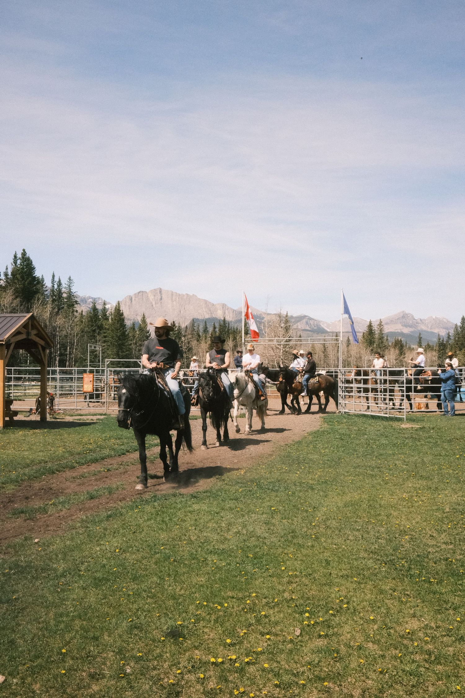 Cowboys riding horses at a rodeo arena with Canadian flags, pine trees, and Rocky Mountains in the background.