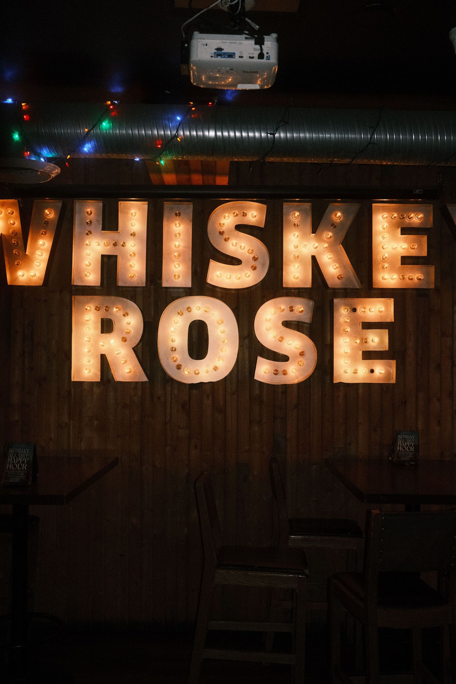 Illuminated marquee letters spelling WHISKEY ROSE on a wooden wall in a dimly lit bar with string lights.