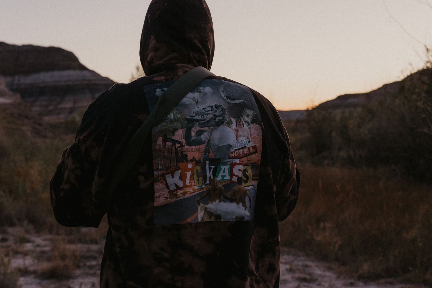 Person wearing camo hoodie with graphic print outdoors at dusk with desert landscape in background.