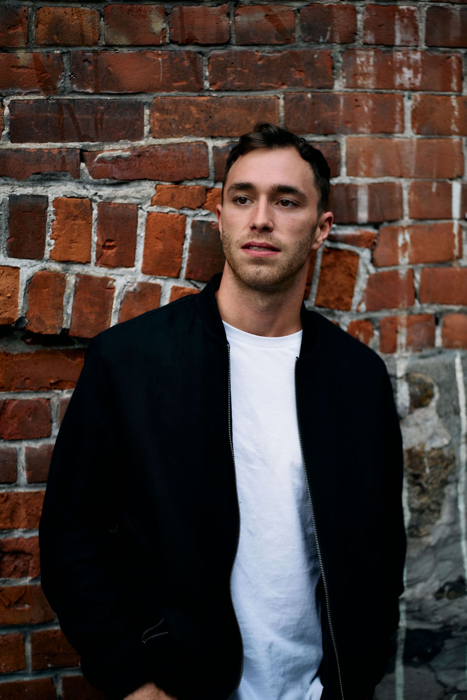 Person in black jacket and white shirt leaning against brick wall in urban setting.