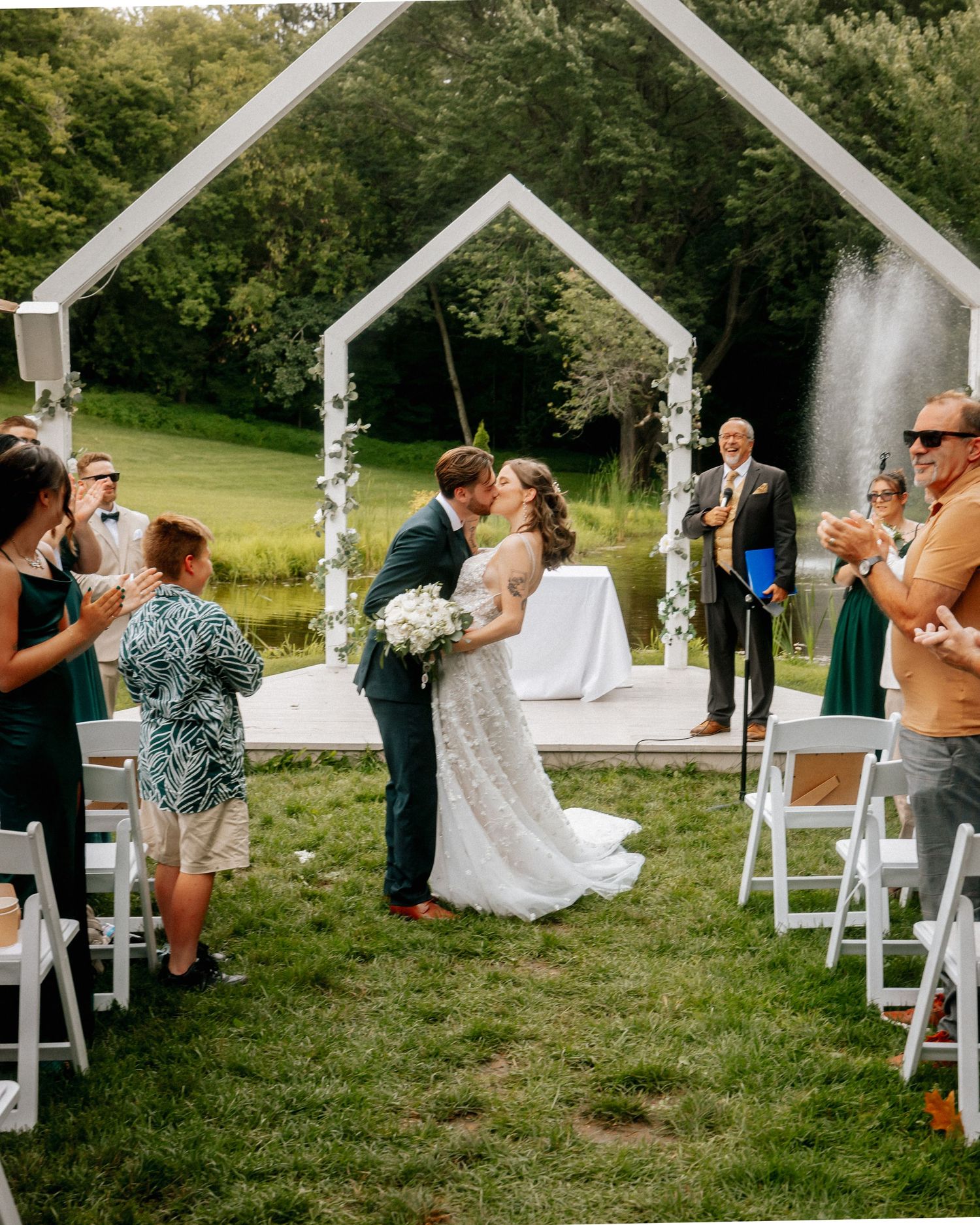 Outdoor wedding ceremony under white arch with guests seated on white chairs.