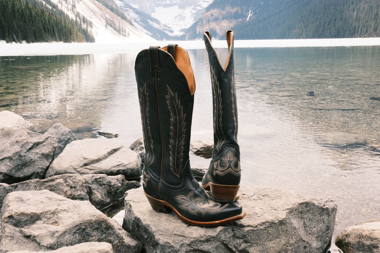 Black cowboy boots standing on rocky shore beside a serene mountain lake with snow-capped peaks in background.