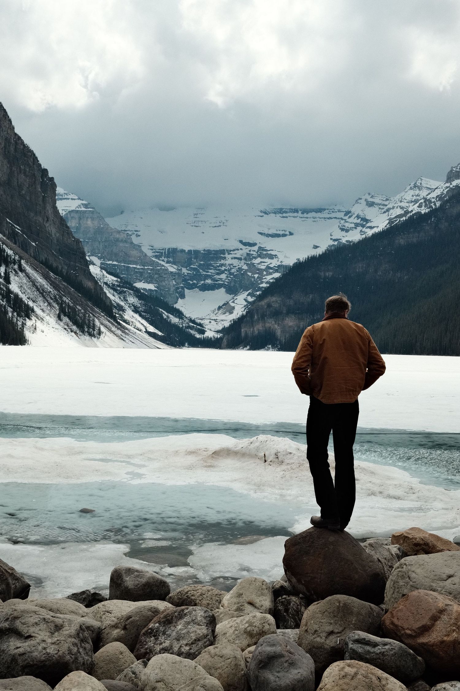 A man in a brown jacket stands on rocks overlooking a partially frozen Lake Louise with snow-capped mountains in winter.