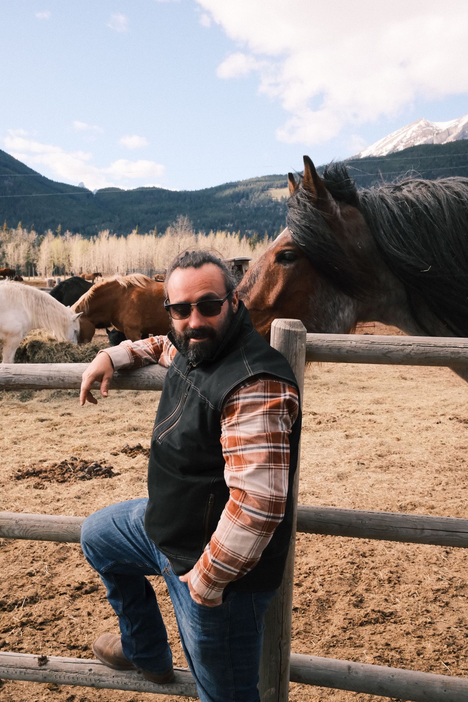Bearded man in sunglasses leaning on wooden fence at horse ranch with mountains in background.