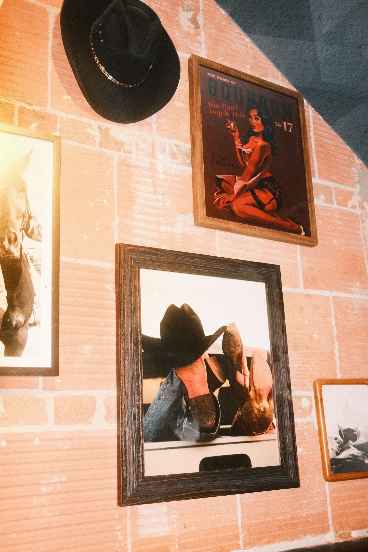 Rustic brick wall display with framed cowboy-themed artwork, a black hat, and vintage magazine cover in warm lighting.