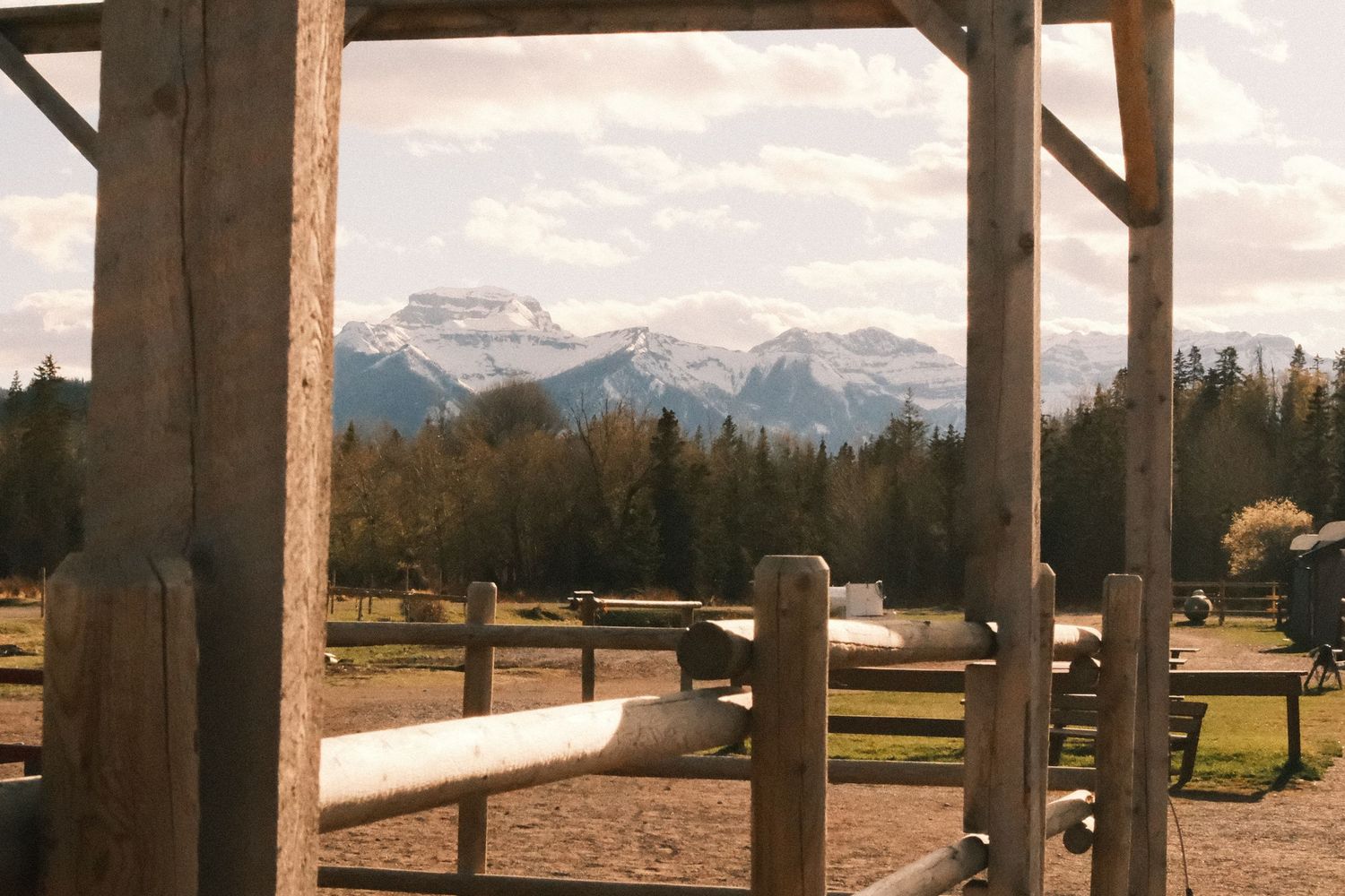 Rustic wooden ranch fence frames a scenic mountain landscape with snow-capped peaks and evergreen trees in the background.