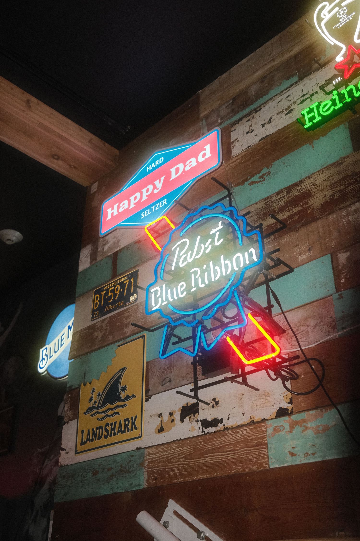 Neon bar signs including Pabst Blue Ribbon and Happy Dad on a rustic multicolored wood plank wall.