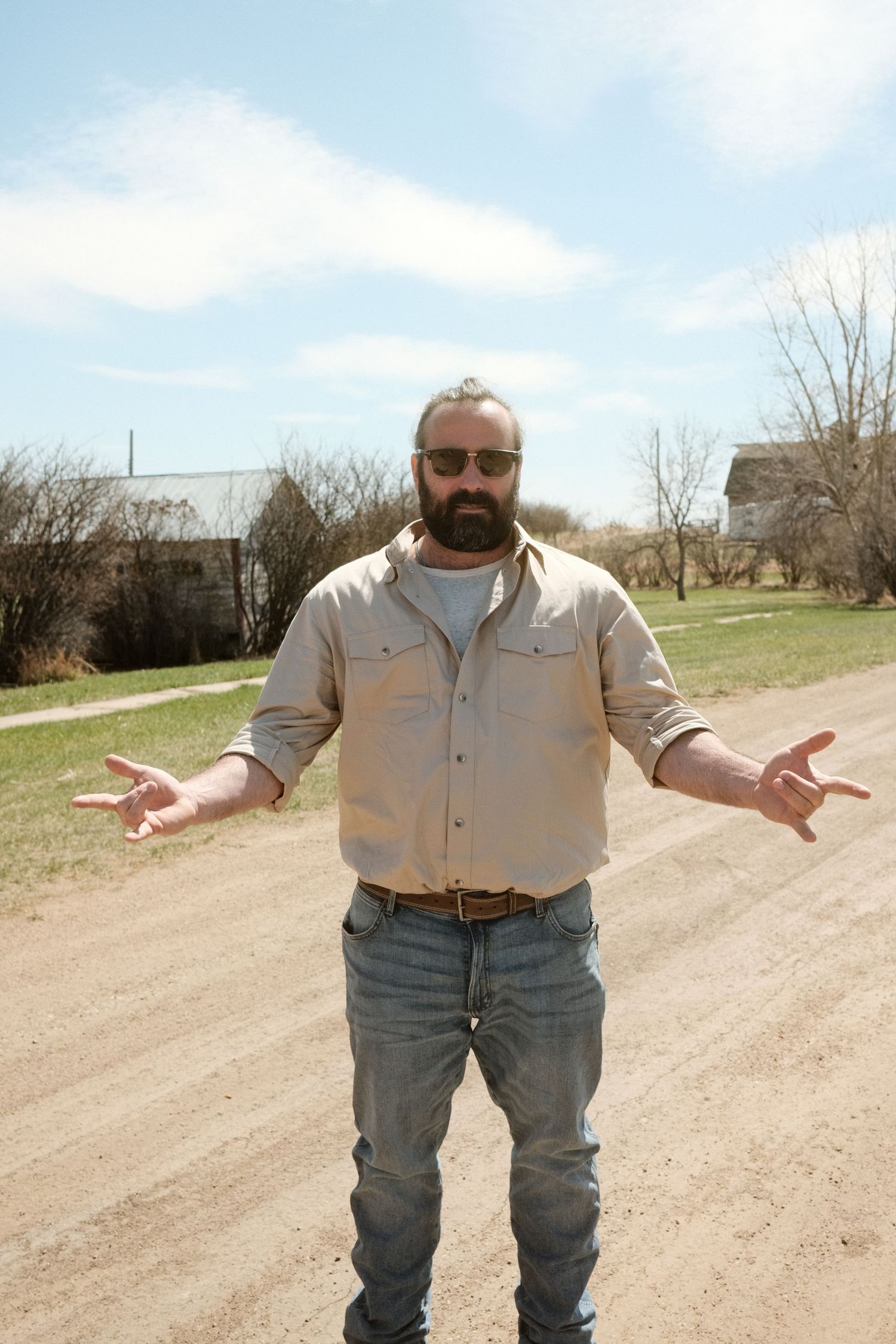 Bearded man in sunglasses and jeans shrugging on a rural dirt road with houses and bare trees in background.