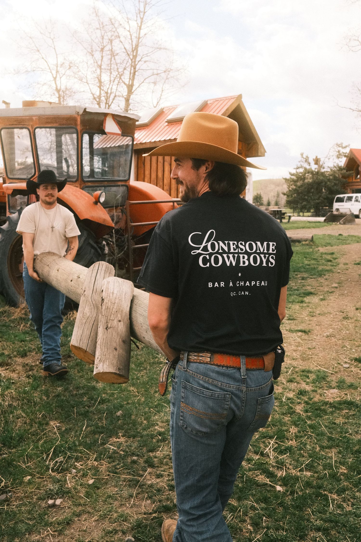 Two cowboys in cowboy hats carry logs near a tractor, one wearing a Lonesome Cowboys Bar a Chapeau black t-shirt.