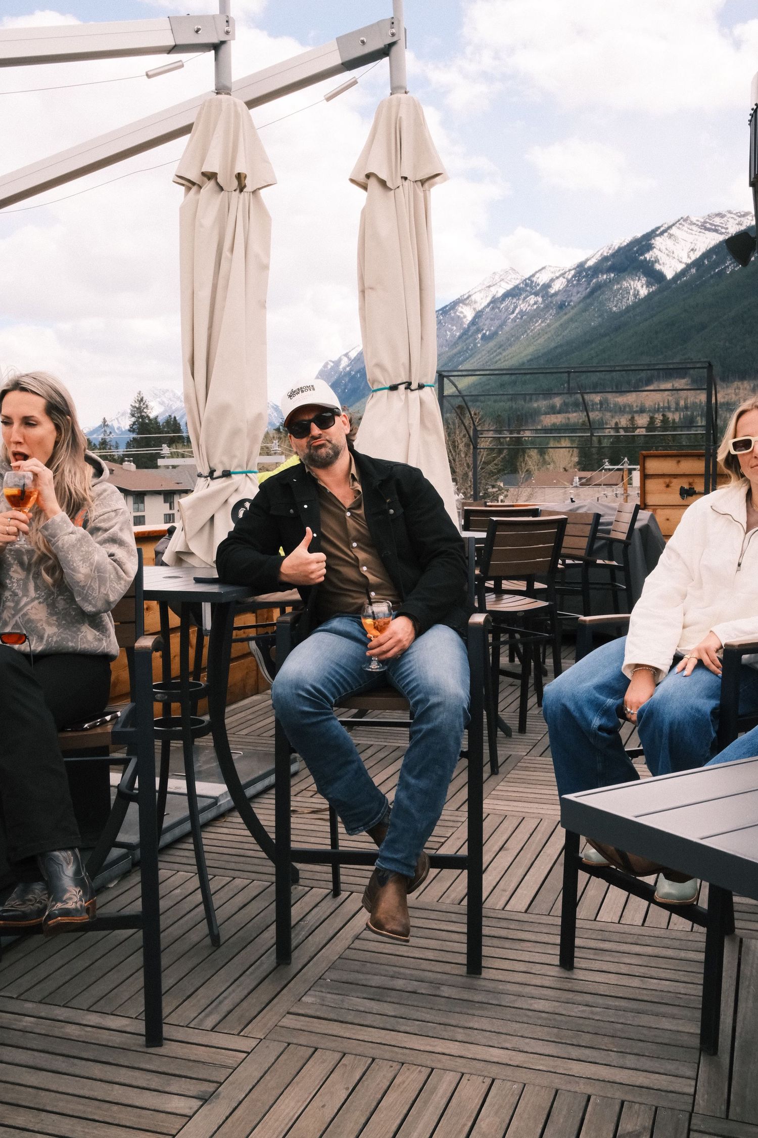 Man in white cap and sunglasses sitting on outdoor patio with mountain views, holding a drink.