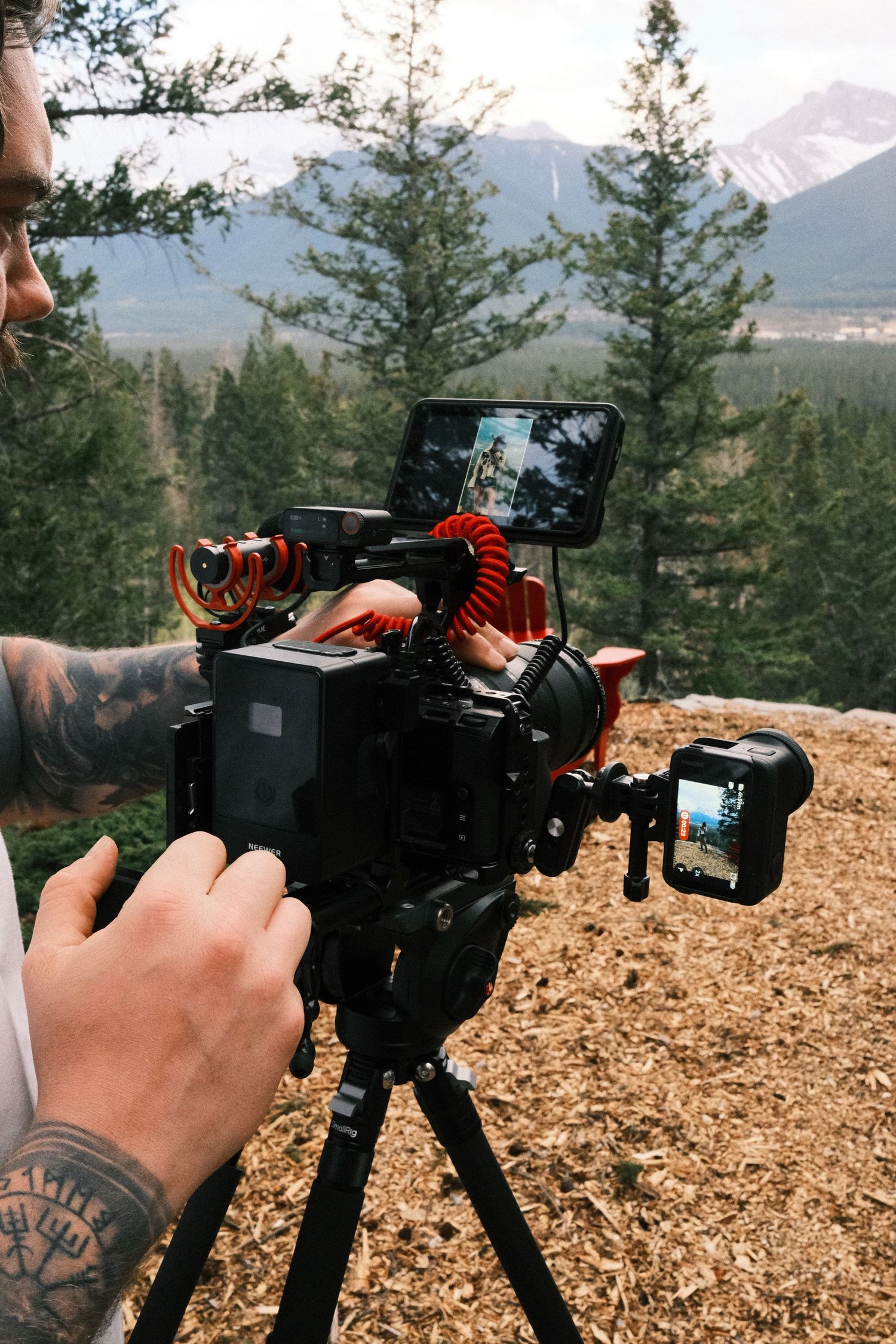 Tattooed videographer operating a professional camera rig on tripod with mountain forest backdrop.