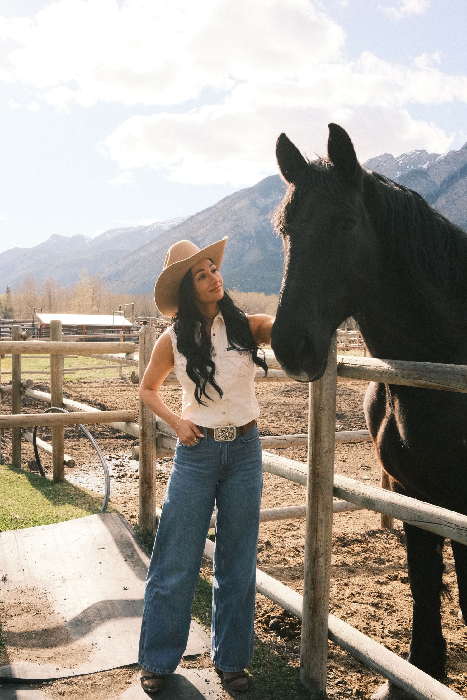 Woman in cowboy hat and jeans standing beside a black horse at a ranch with mountain backdrop.